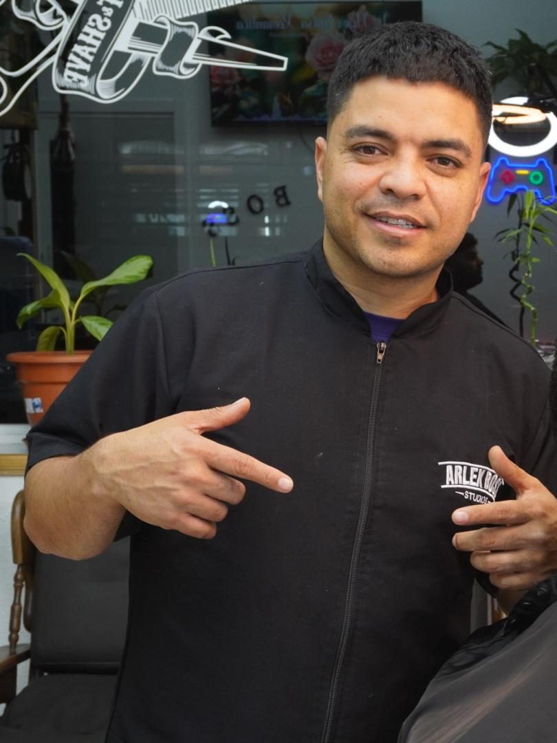 Arlex en su barbería, señalando el logotipo de Arlex Boss Studios